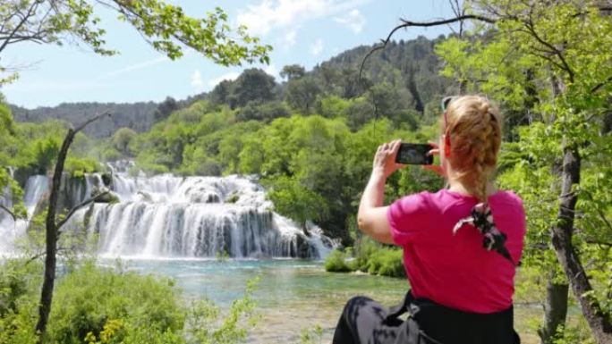 金发碧眼的女人坐在Krka国家公园Skradin瀑布前的一块岩石上，用智能手机拍照