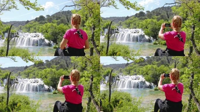 金发碧眼的女人坐在Krka国家公园Skradin瀑布前的一块岩石上，用智能手机拍照