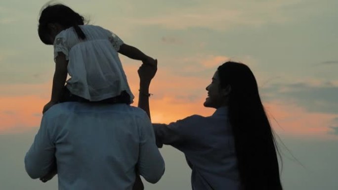 后视图，4岁的亚洲女婴剪影家庭骑着父亲的脖子，母亲牵着她的手，看着海滩上的日落。家庭假期快乐。伸出援