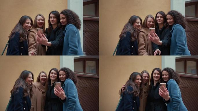 一群美丽的多民族快乐的年轻女性在米色建筑使用智能手机一起微笑自拍照片。