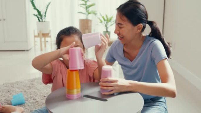 亚洲蹒跚学步的小女孩和妈妈坐在地毯上，开心的时刻在家里客厅的桌子上玩彩色堆栈杯玩具。