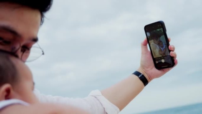 周末在海滩上旅行时，特写父亲抱着新生婴儿并使用手机自拍