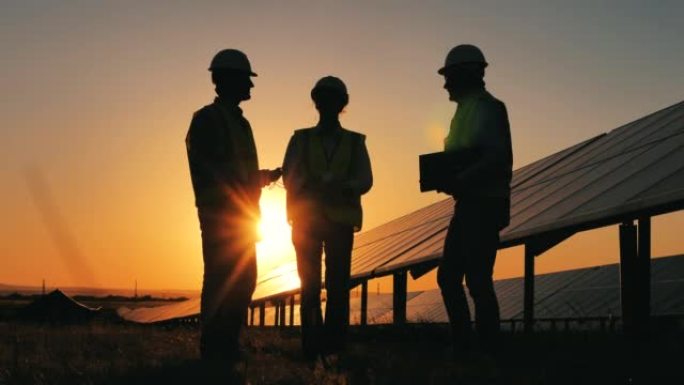 日落时，背光工程师正在太阳能电池板旁边进行讨论。可再生能源、太阳能发电厂、绿色电力概念。