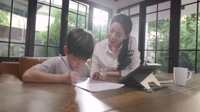 一个亚洲男孩正坐在家里专心地在线学习，记下他通过平板电脑学到的知识，母亲在他身边教书。由于Covid