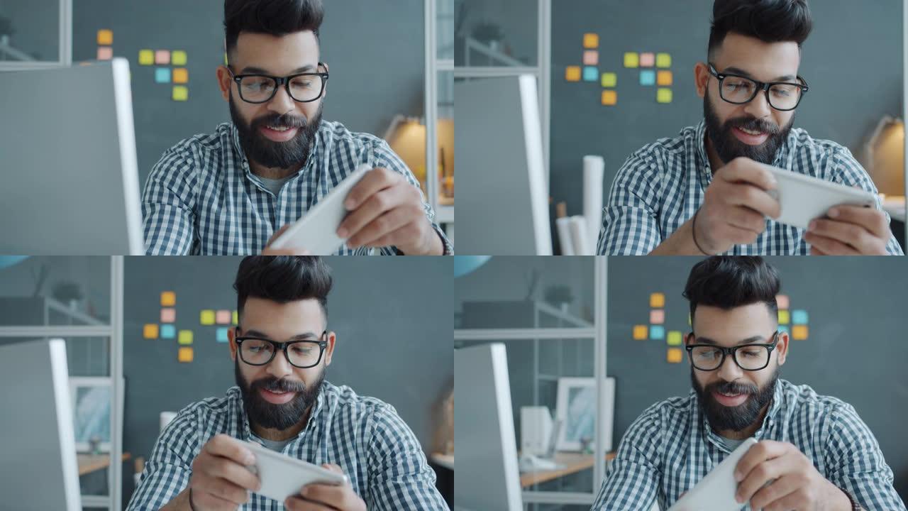 微笑的阿拉伯人独自享受在工作场所玩智能手机视频游戏