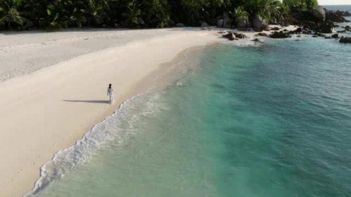 一名妇女在热带岛屿上的碧绿海水中沿着原始海滩的水边行走的空中后视