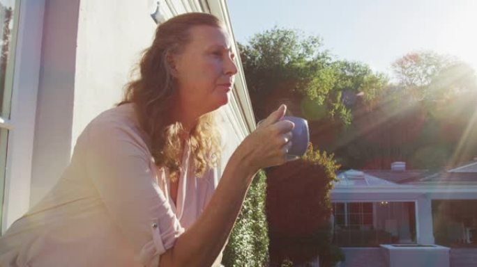 体贴的白人高级妇女站在阳光明媚的阳台上喝咖啡，环顾四周