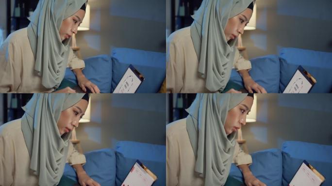 特写迷人的年轻亚洲穆斯林美女戴着头巾，随意坐在沙发上，晚上在家舒适的客厅使用笔记本电脑远程工作。女孩