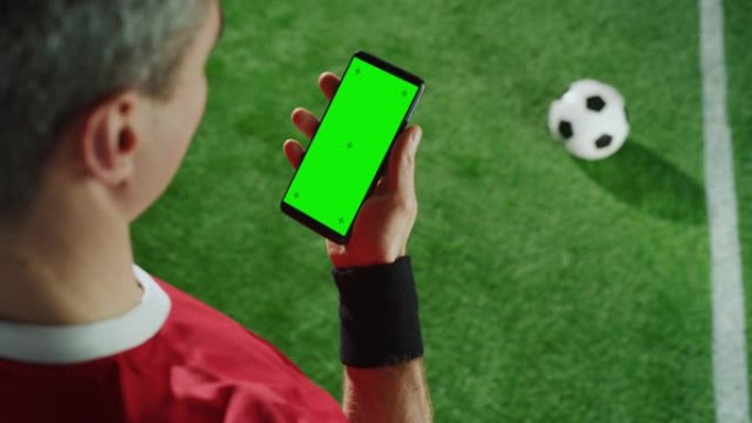 职业足球运动员持有智能手机与绿色屏幕色度键跟踪标记。足球明星站在球场上使用互联网时，从上到下的肩部拍
