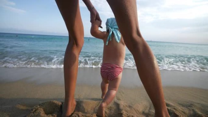 一位年轻母亲的真实特写镜头正在帮助她蹒跚学步的男婴迈出第一步，并在阳光明媚的日子里教她在海边的沙滩上