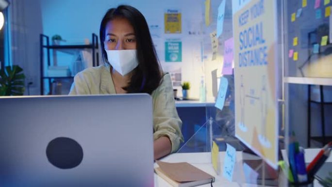 快乐亚洲女商人戴着医用口罩，在新的正常情况下保持社交距离，以预防病毒，同时在办公室晚上工作时使用笔记