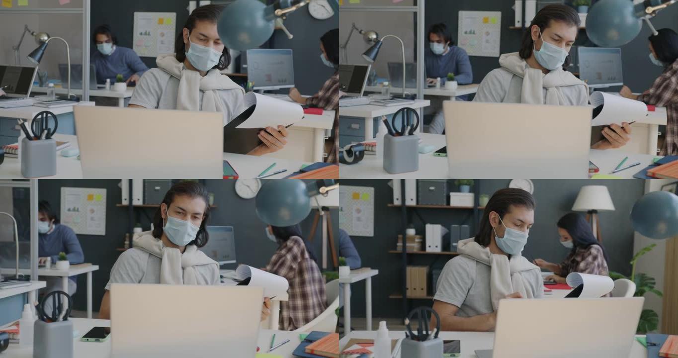 男性员工戴着医用口罩在办公室工作，使用笔记本电脑和检查文件