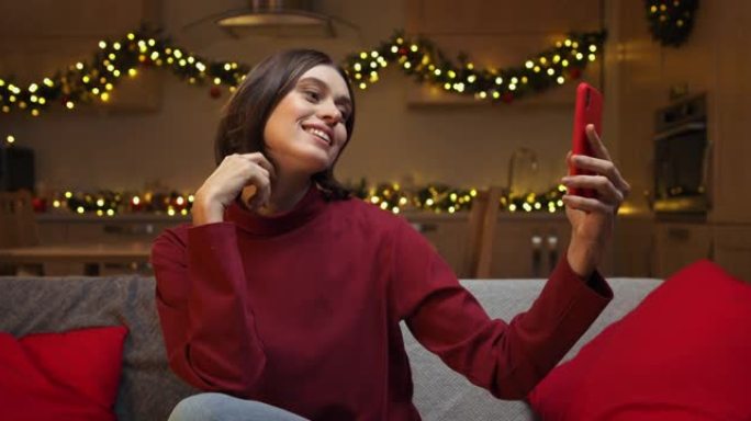 一位极具吸引力的棕色头发女人坐在舒适的圣诞节装饰房间的沙发上，在她的红色智能手机上自拍，笑容灿烂