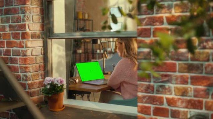 女性坐在时尚客厅的桌子后面，在带有绿屏模拟显示的笔记本电脑上工作。业务经理在家远程工作。从窗外看。