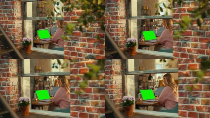 女性坐在时尚客厅的桌子后面，在带有绿屏模拟显示的笔记本电脑上工作。业务经理在家远程工作。从窗外看。