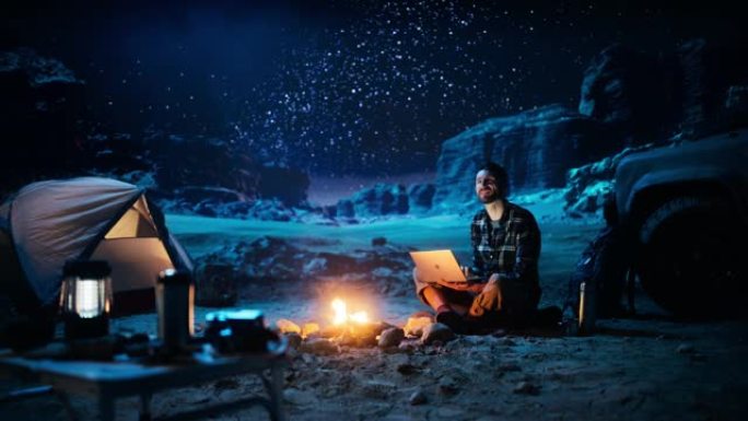 坐在篝火旁的男性旅行者在峡谷中露营时使用笔记本电脑。在度假旅行时通过互联网进行数字远程工作，电子商务