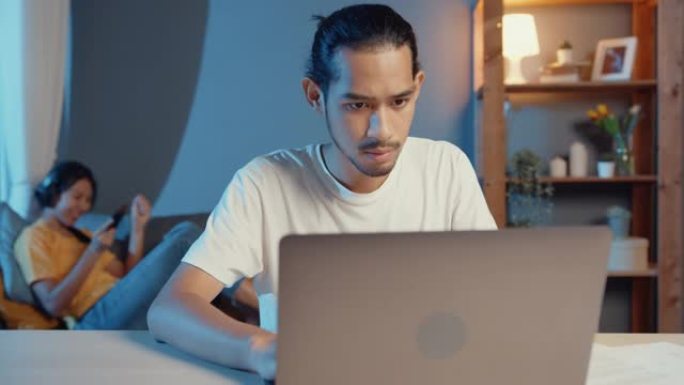 自由职业者亚洲夫妇在休闲丈夫的焦点工作在笔记本电脑晚上妻子休息一下，感觉放松戴耳机在智能手机上播放音