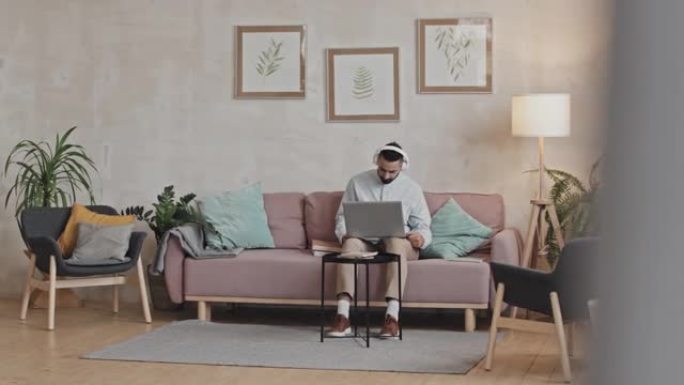 戴着耳机的男人在家中沙发上使用笔记本电脑