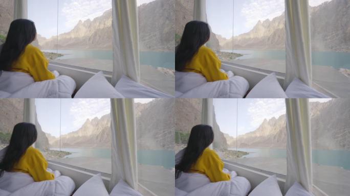 早晨醒来并透过湖边的窗户看时，穿着黄色衣服并包裹在温暖的毯子中的年轻女子的后视图。种族多样化的旅行者