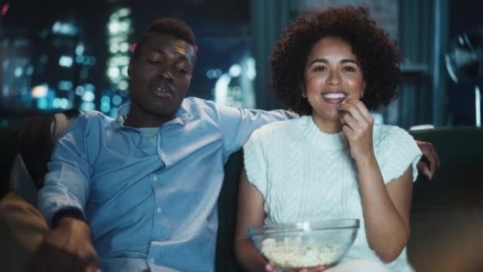 黑人夫妇在电视上看喜剧电影，深夜坐在公寓的沙发上吃爆米花。笑男女朋友在家一起欣赏搞笑电视剧。肖像照