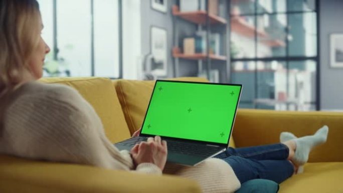 美丽的高加索专家躺在沙发上，在家里的客厅用绿屏模拟显示笔记本电脑工作。自由女性在社交网络上通过互联网
