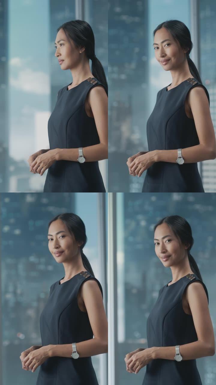 一位亚洲女商人的垂直肖像，穿着时髦的黑色连衣裙，使用笔记本电脑，在城市办公室的窗户旁边摆姿势。自信的