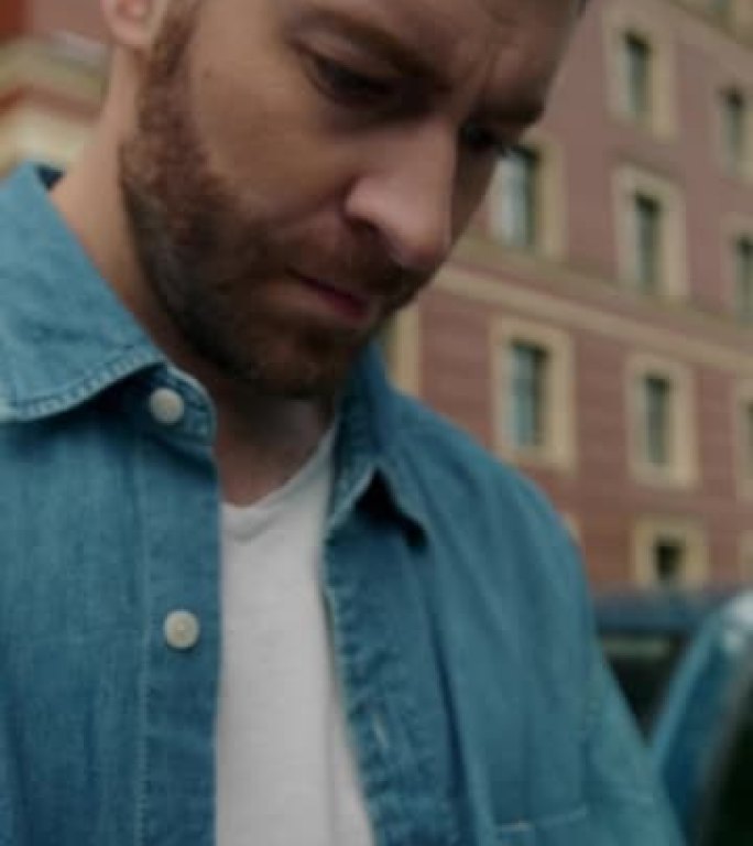 一个穿着牛仔裤衬衫的男人的特写镜头，他靠在一辆蓝色的汽车上，在手机里打字