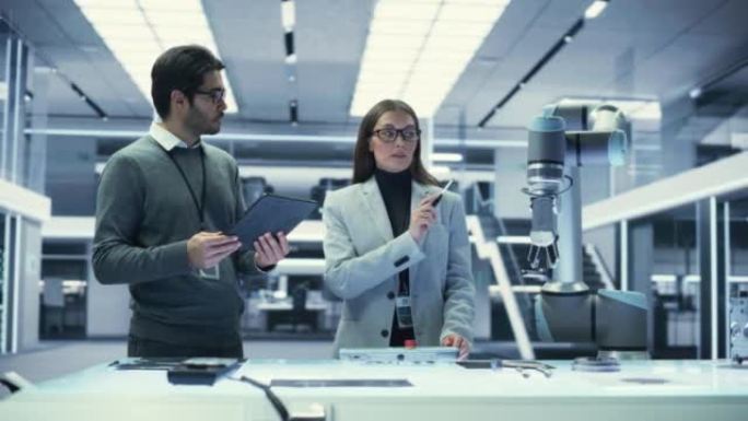 两名工业机器人工程师用机械臂聚集在一张桌子旁。女性和男性科学家使用平板电脑来操纵和编程机器人，以拾取