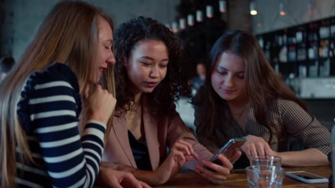 友谊和团结。三个年轻美丽快乐的多种族女性在咖啡店的桌子上一起使用智能手机。