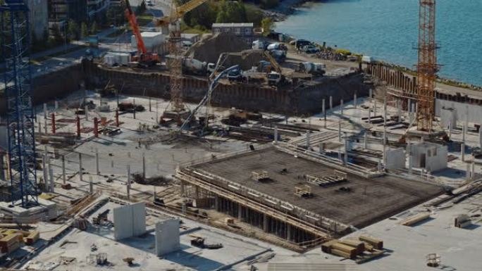 高空飞行在新的建筑开发场地上，用高塔起重机建造房地产。雇用重型机械和建筑工人。背景中的蓝色海洋和城市