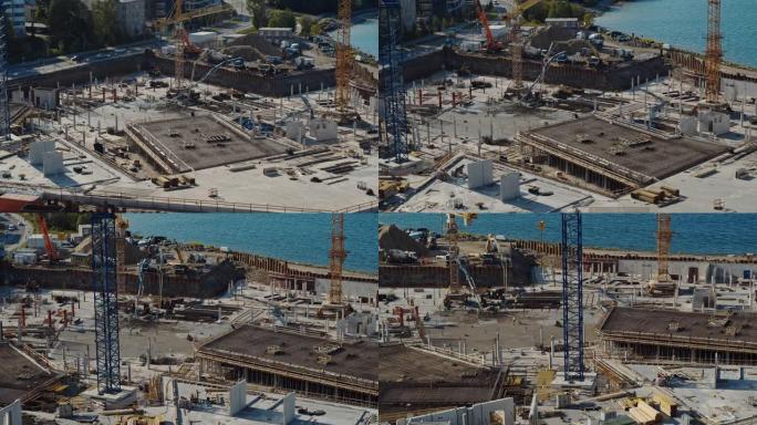 高空飞行在新的建筑开发场地上，用高塔起重机建造房地产。雇用重型机械和建筑工人。背景中的蓝色海洋和城市
