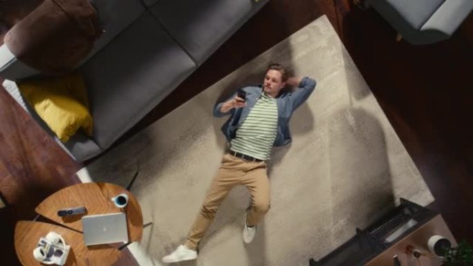 公寓:英俊的男人躺在客厅的地板上，用智能手机。男人在地毯上放松。自由职业者远程工作，电子商务，网上购