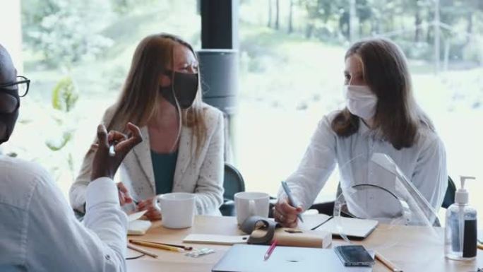 与大流行安全措施的商务会议。年轻快乐的多种族同事戴着口罩在办公室的桌子上交谈。