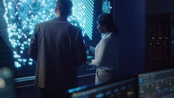 项目经理和计算机科学工程师在使用大屏幕显示神经连接和数据时交谈。电信公司系统控制和监控室。