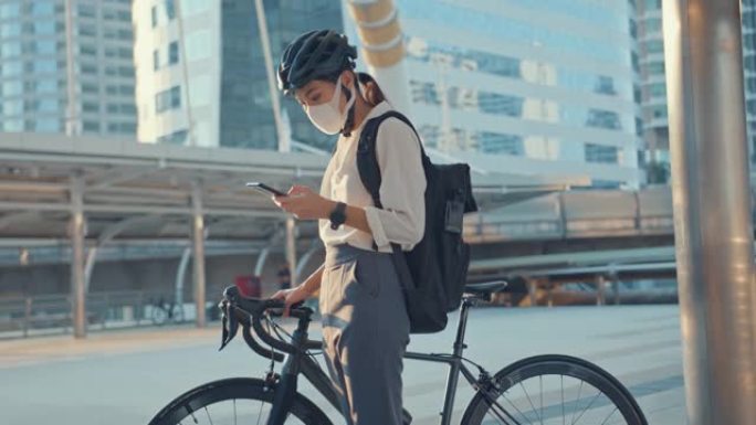 亚洲女商人背着背包戴防病毒保护面具骑自行车步行并在城市街道检查电话去办公室上班。