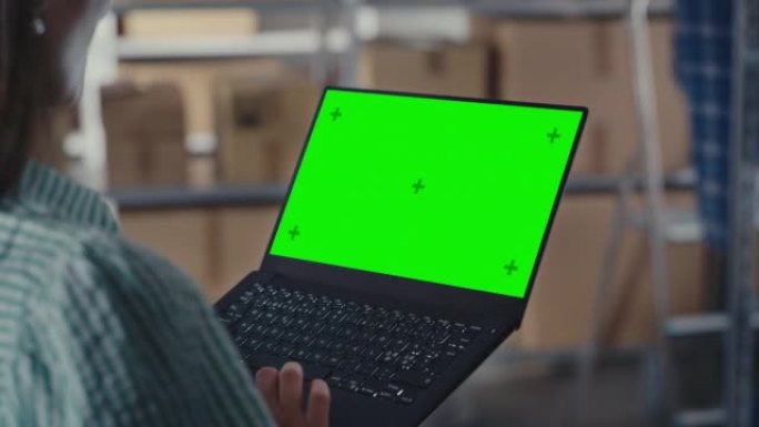 女人使用带有绿屏Chromakey模拟显示的笔记本电脑。员工浏览互联网，在带有包裹的在线商店仓库储藏