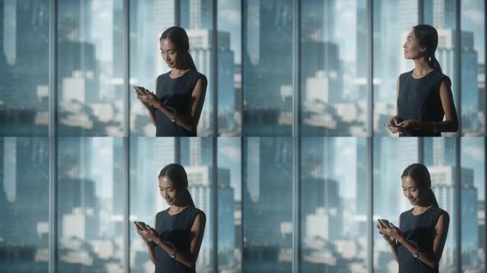 成功的女商人穿着时髦的衣服站在现代办公室，使用智能手机，看着窗外的大城市的摩天大楼。自信的从事金融项
