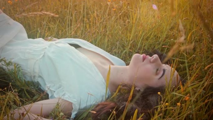 日落时分，穿着白色连衣裙的SLO MO年轻女子躺在草地上