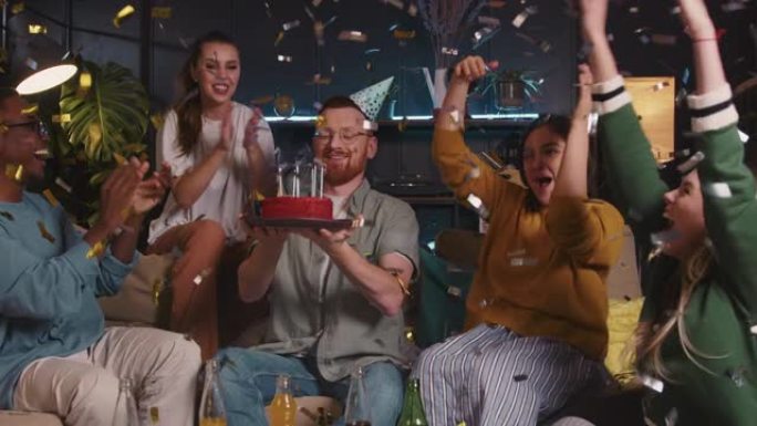 生日蛋糕快乐微笑的红发男人在与多民族朋友庆祝的有趣五彩纸屑派对上吹蛋糕。