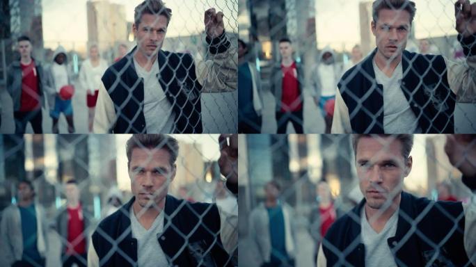 一个英俊的白人男子的肖像，他拿着栅栏，摆姿势，看着相机，同时站在城市的户外。站在他身后的时尚多元朋友