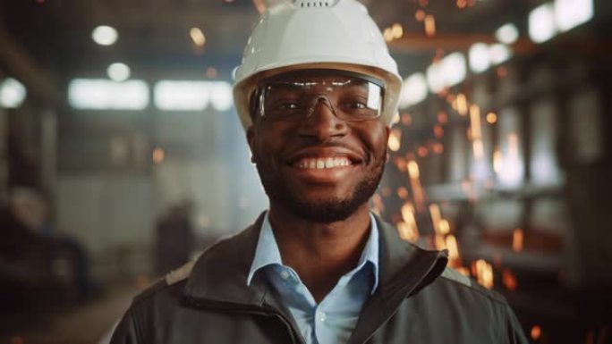 快乐专业的重工业工程师/工人，穿着制服，戴着眼镜，戴着安全帽在钢铁厂。微笑的非洲裔美国工业专家站在金