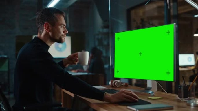 年轻英俊的专家在创意办公室的台式电脑上工作，绿屏模拟显示。男经理从他从工作台上拿起的白色杯子里喝了一
