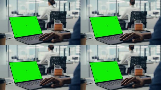 成功的黑人商人坐在办公桌前，在办公室里用绿屏笔记本电脑工作。使用色度键显示的匿名商人。时尚明亮的工作