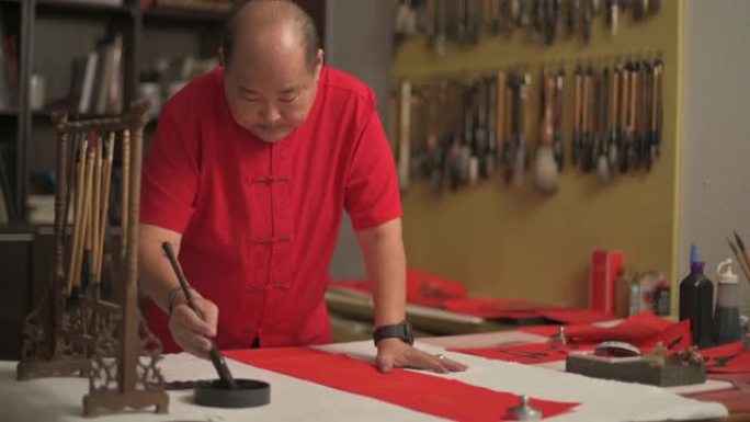 亚洲中国男性为即将到来的农历新年庆祝活动而练习中国书法，目的是通过将其写在红色纸上来实现繁荣和良好的