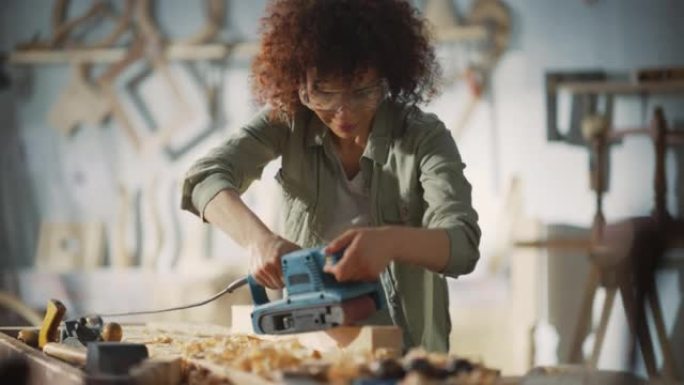 多种族的女木匠戴着防护安全眼镜，并使用电动皮带砂光机研磨木块。艺术家或家具设计师在车间从事产品创意。