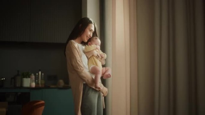 美丽的年轻亚洲女子怀里抱着她的孩子，站在家里的窗户旁边。可爱的小孩在母亲的怀抱中休息，因为她向他展示