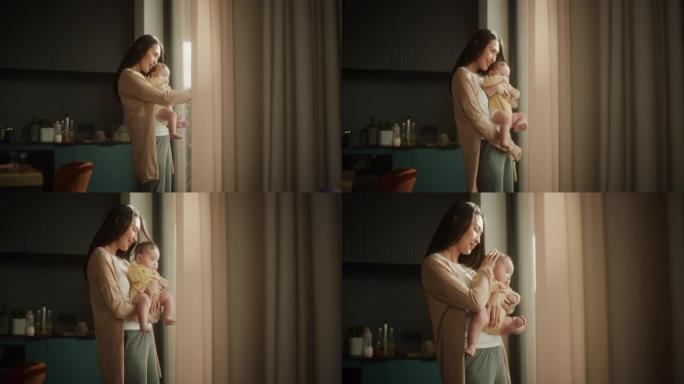 美丽的年轻亚洲女子怀里抱着她的孩子，站在家里的窗户旁边。可爱的小孩在母亲的怀抱中休息，因为她向他展示