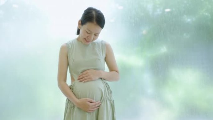 怀孕的亚洲母亲站在花园窗户前揉着肚子。