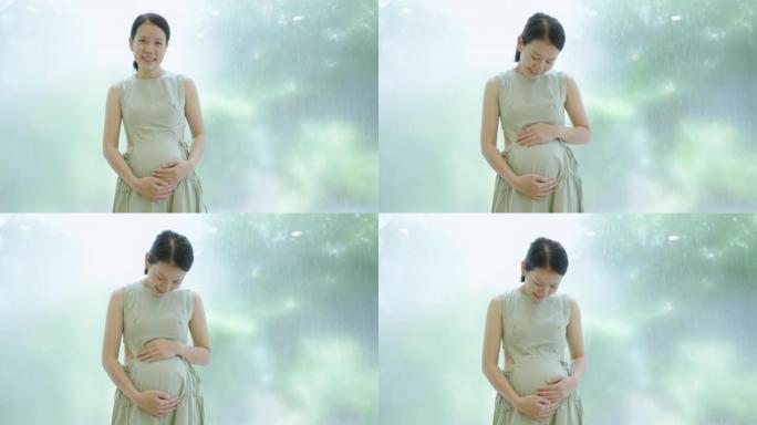 怀孕的亚洲母亲站在花园窗户前揉着肚子。