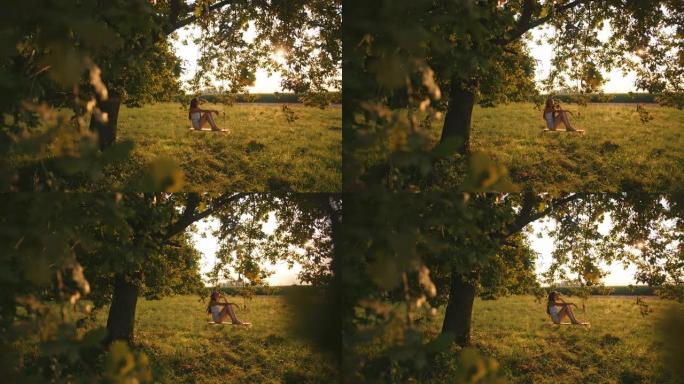 慢动作，手持一个年轻女孩摇摆的镜头，坐在一个悬挂在阳光充足的草地上的大树枝上的绳索秋千上